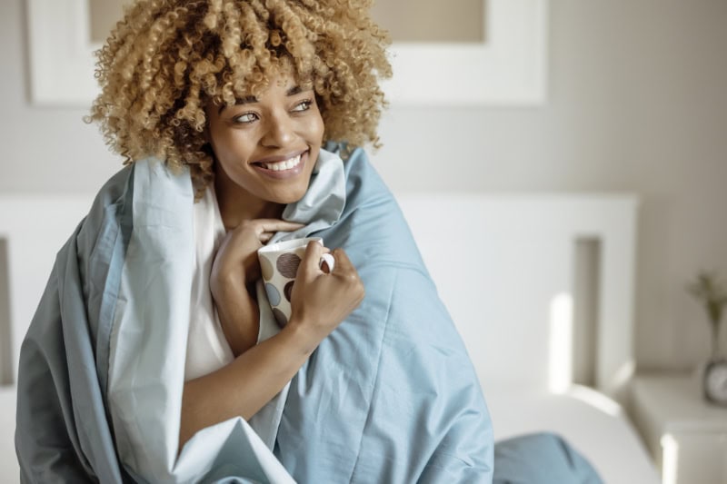 How Do I Keep My Heat During Extreme Cold Weather? Pretty Woman Snuggled Under Duvet With Cup of Coffee or Tea in Bed at Home Bedroom. Morning, Leisure and People Concept